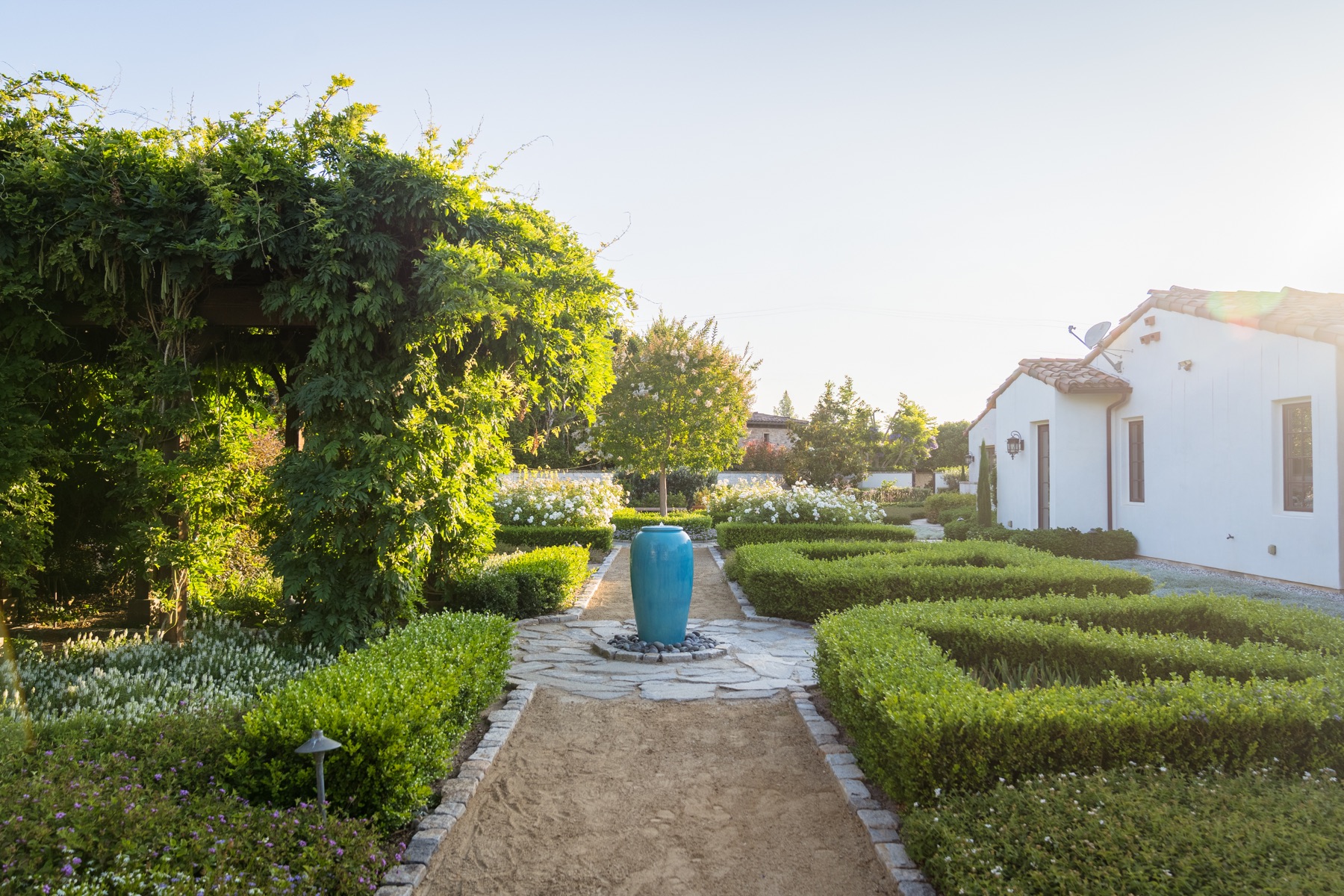 Gravel garden path between clipped boxwood hedges toward a turquoise urn fountain and a white home, landscaped by Evoscapes.