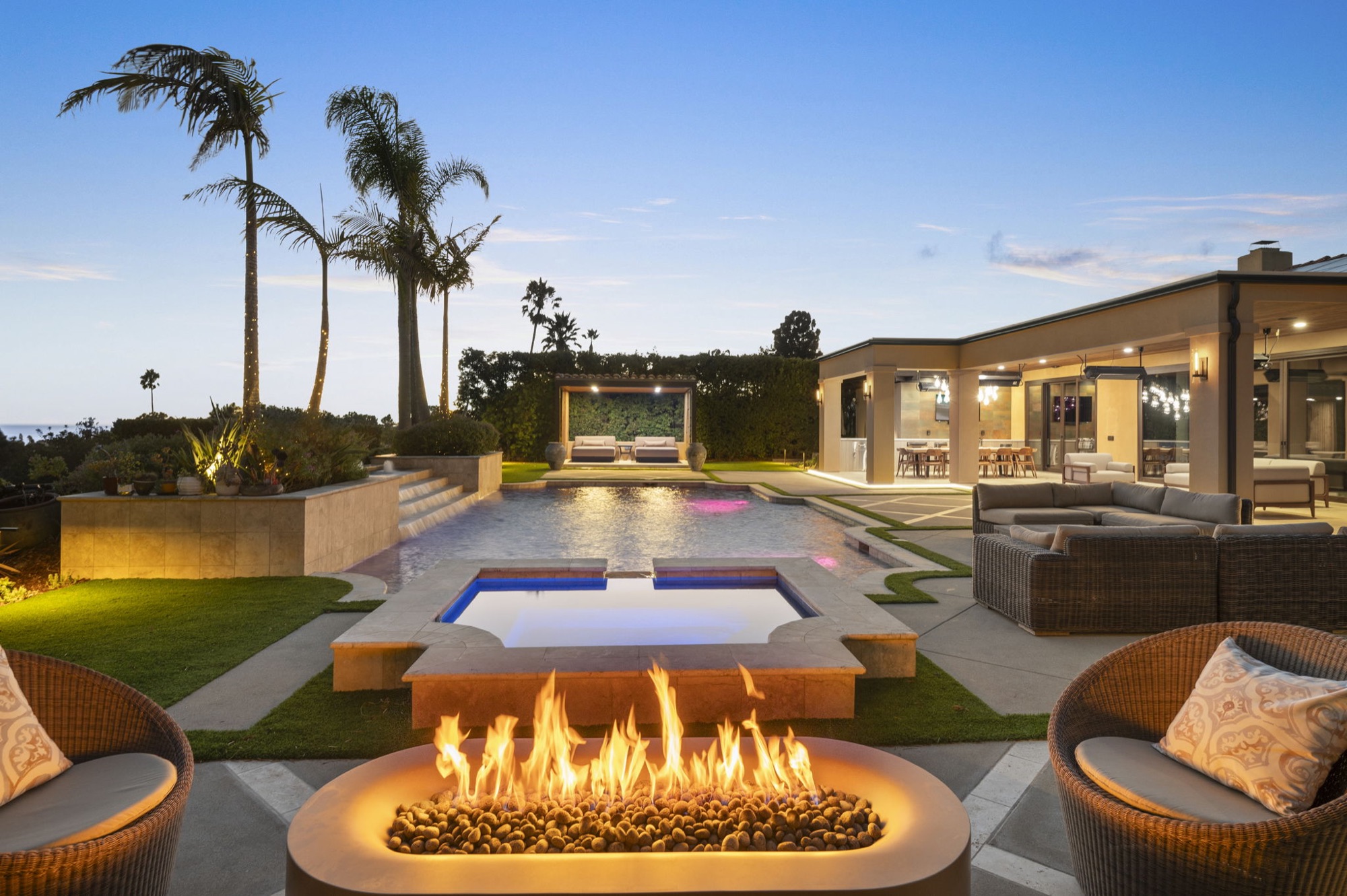Backyard at dusk with a fire feature, pool, palms and an open-sided pavilion, built by Evoscapes.