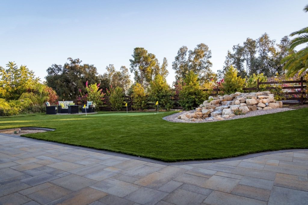 Paver patio edging a turf lawn and putting green with a rock water feature, by Evo Turf and Pavers.
