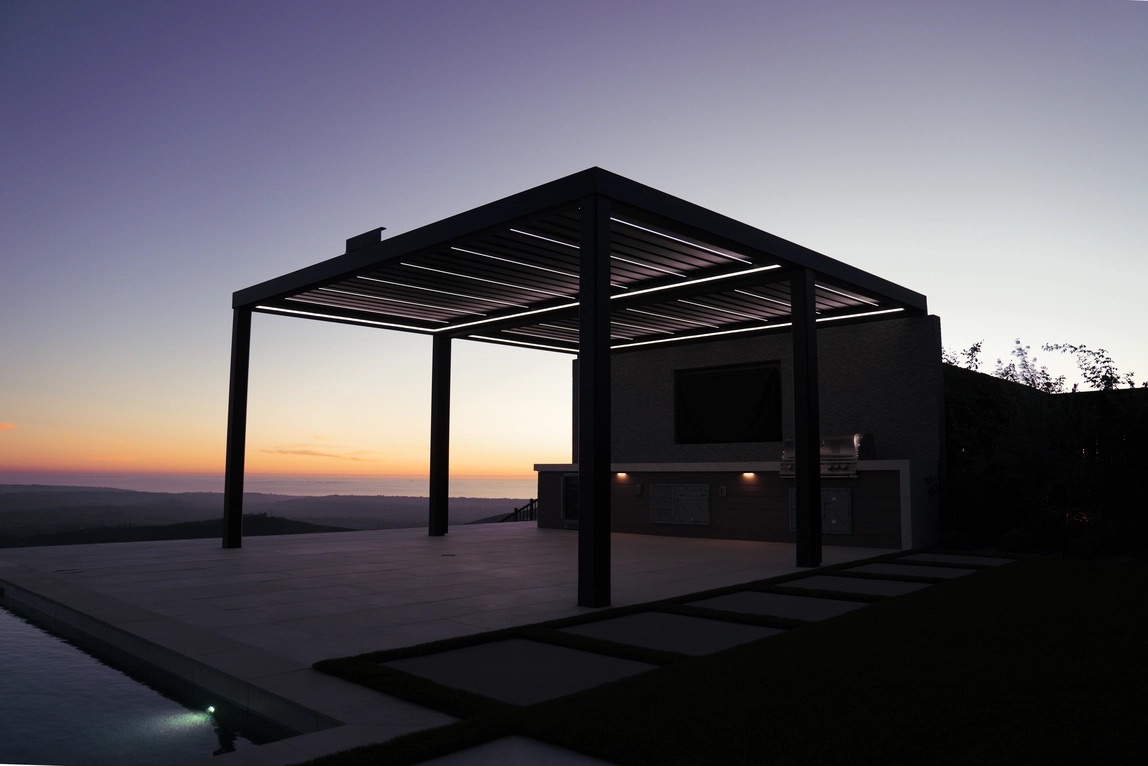 Louvered pergola with illuminated slats over a patio and an outdoor grill at dusk.